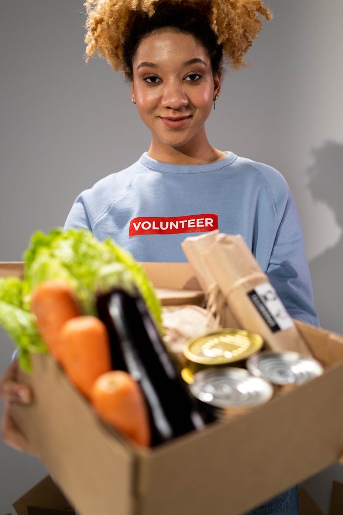 stats-img Smiling woman holding a box filled with various vegetables and canned goods during a volunteer drive.